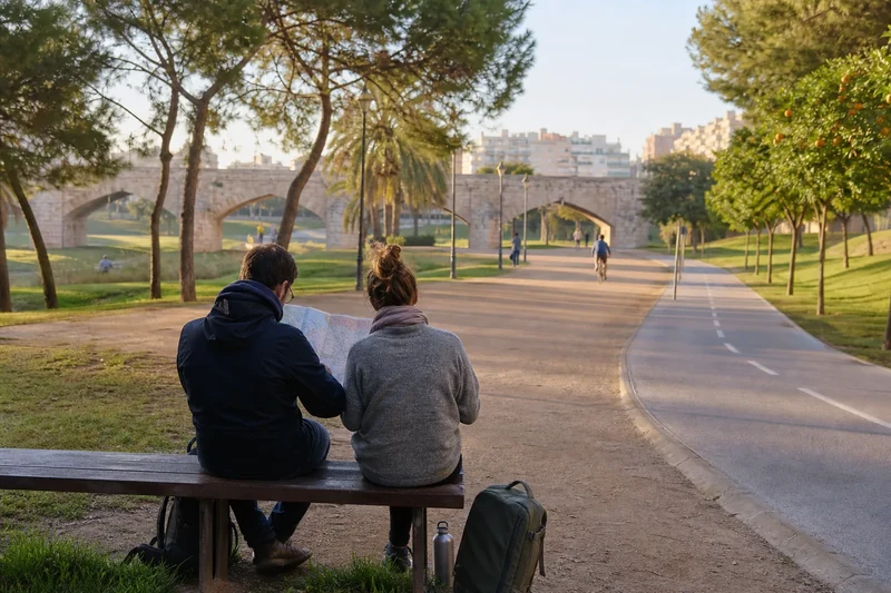 Dos viajeros ficticios consultan un plano en un banco del Jardín del Turia, frente a senderos y un puente de piedra representativo