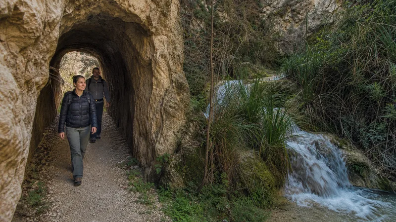 Senderistas atraviesan un túnel rocoso junto al agua en un paisaje inspirado en Chelva