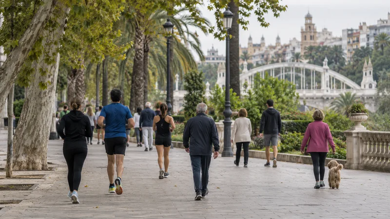 Personas caminando y trotando por una alameda arbolada de València, con un puente blanco al fondo.