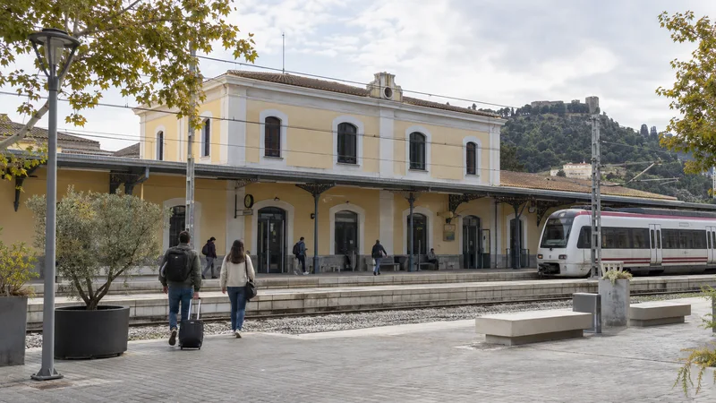 Fachada de una estación ferroviaria con viajeros en el andén y un tren de Cercanías.