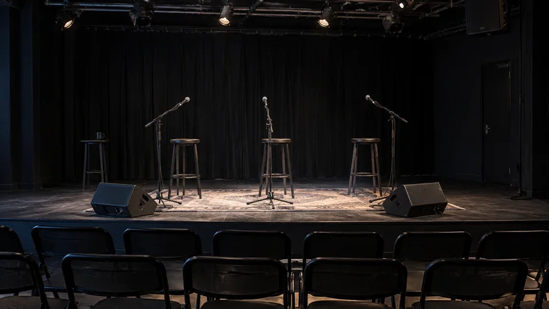 Escenario vacío de teatro en sala negra con tres micrófonos, cuatro taburetes y varias filas de butacas.